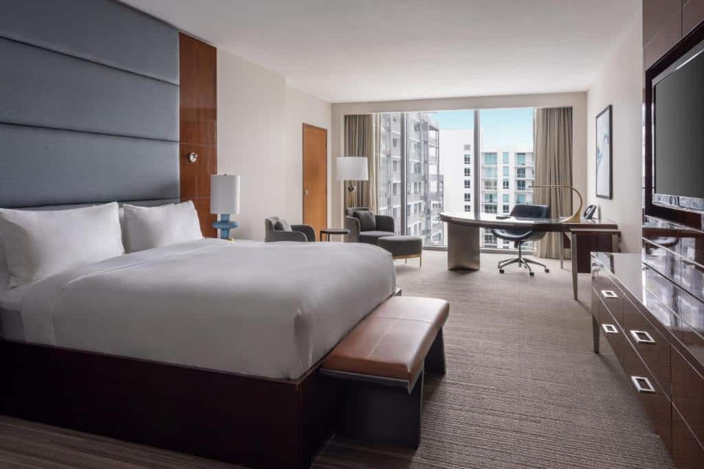 Imagem do quarto do JW Marriott Marquis Miami com cama de casal do lado esquerdo da imagem com um banco no pé da cama do lado esquerdo uma cômoda com luminária em frente a cama uma cômoda com TV presa na parede do lado esquerdo da cama ao fundo uma pequena sala de estar com mesa e cadeira. Representa hotéis 5 Estrelas em Miami.