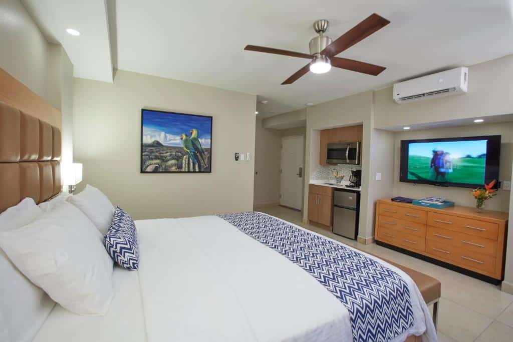 Quarto do All Inclusive - Divi Dutch Village Beach Resort. Uma cama de casal do lado esquerdo, de frente uma cômoda com televisão e ar-condicionado. Ao lado uma minicozinha e a porta do quarto. No meio, em cima, um ventilador.