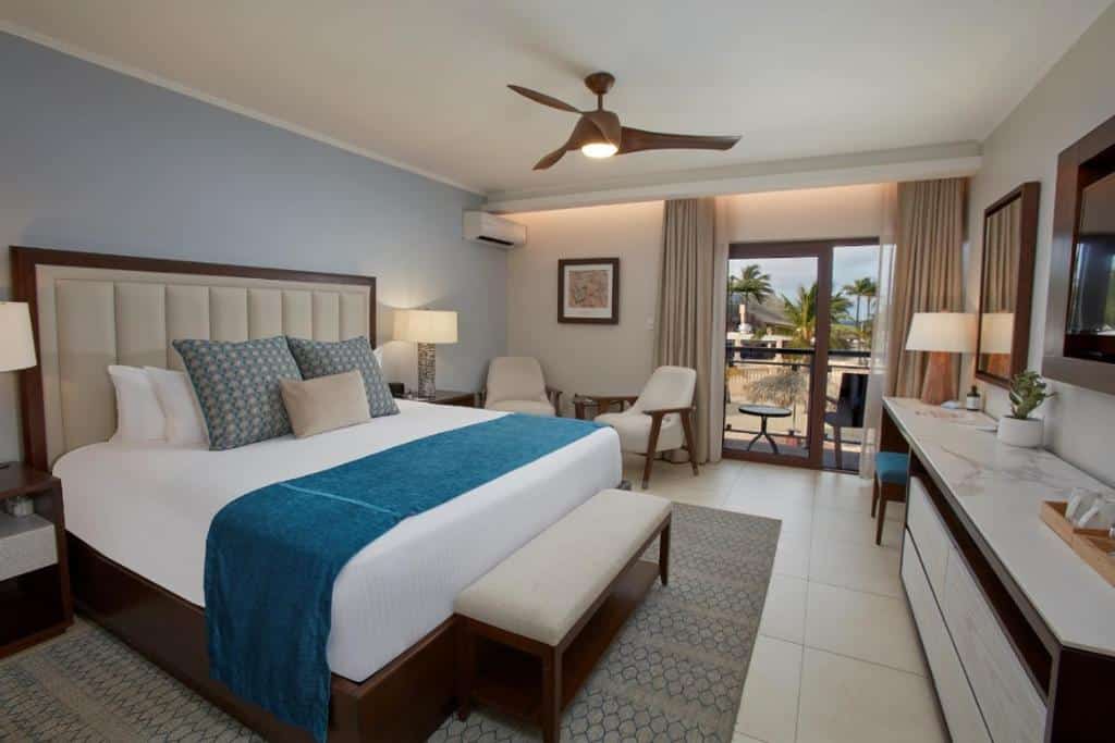 Quarto do Manchebo Beach Resort and Spa. Uma cama de casal do lado esquerdo, com uma cômoda e um abajur de cada lado. No fundo duas poltronas e um ar-condicionado. Do lado direito uma raque, no fundo um abajur e a porta da varanda.