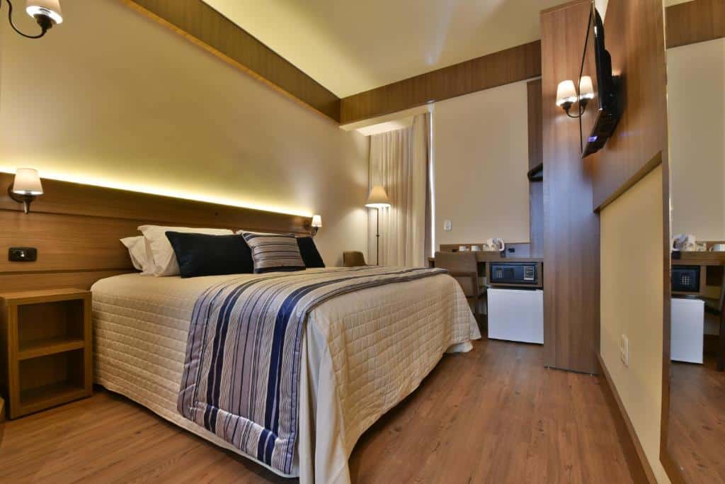 Quarto do Hotel Due Nobili. Uma cama de casal do lado esquerdo, com uma cômoda e uma luminária de cada lado. Do lado direito um espelho, uma televisão e uma luminária. No fundo, uma mesa de trabalho, um abajur, um cofre e um frigobar. Foto para ilustrar post sobre hotéis baratos em Gramado.