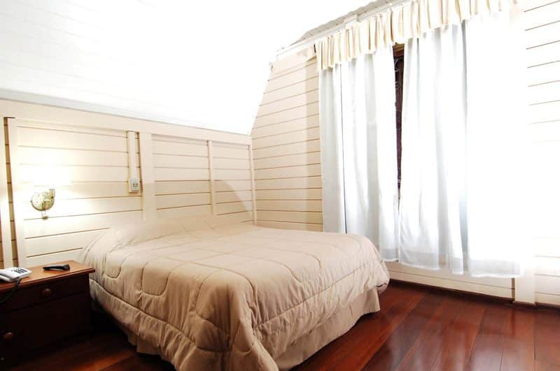 Quarto do Hotel Pousada das Flores. Do lado direito cortina tampando a janela, no lado esquerdo uma cama de casal e uma cômoda com telefone e controle. Foto para ilustrar post sobre hotéis baratos em Gramado.
