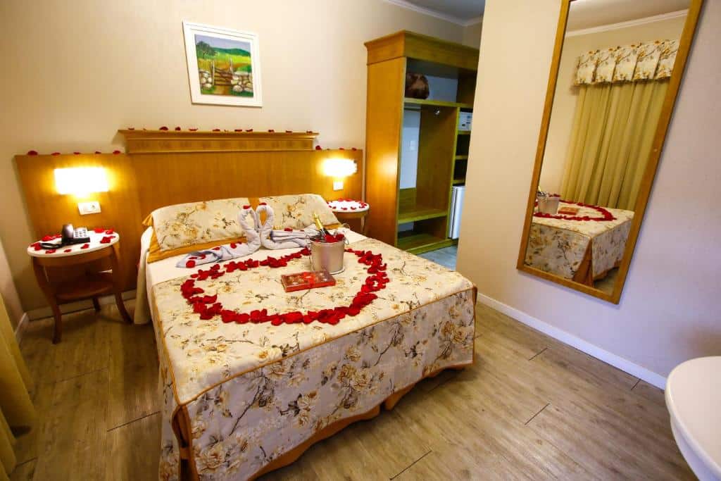 No meio uma cama de casal do Life Hotel Infinity. Uma cama toda decorada com pétalas, chocolate e champagne. De cada lado uma mesa com abajur. No lado direito, um espelho e um corredor com guarda-roupa e frigobar. Foto para ilustrar post sobre hotéis baratos em Gramado.