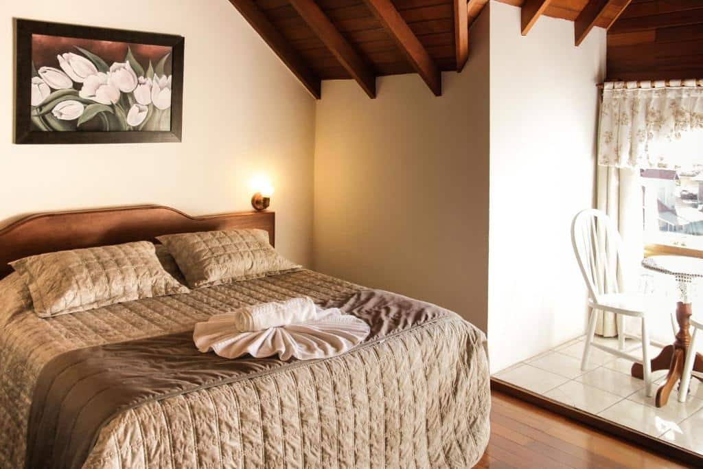 Quarto do Hotel Aconchego da Serra. Uma mesa com duas cadeiras no lado direito. No lado esquerdo uma cama de casal. Foto para ilustrar post sobre hotéis baratos em Gramado.
