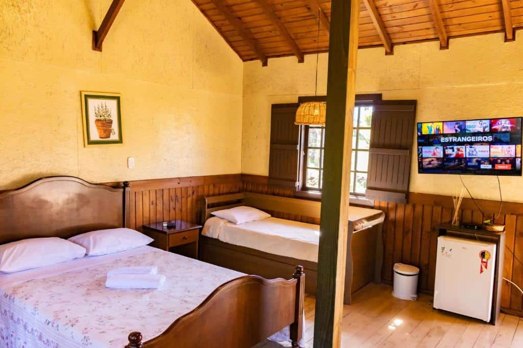Quarto da Pousada Villa da Uva. Uma cama de casal e uma cama de solteiro do lado esquerdo, em cima, uma janela. No lado direito uma televisão, em baixo um frigobar e um lixo. Foto para ilustrar post sobre hotéis baratos em Gramado.