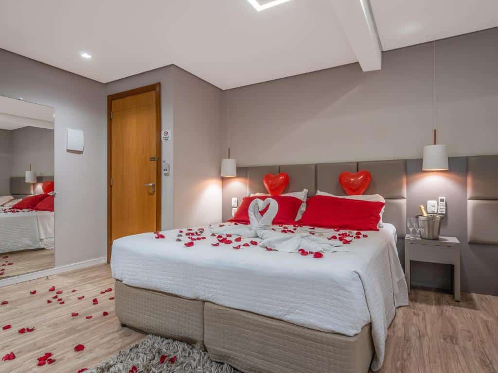 Quarto da Alameda Felice Hotel. Uma cômoda com um champagne e duas taças do lado direito, no meio, uma cama de casal com pétalas e corações em cima, de cada lado um lustre. No lado esquerdo, um espelho e uma porta do quarto. Foto para ilustrar post sobre hotéis baratos em Gramado.