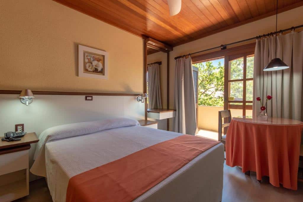 Quarto do Residencial Vale Dourado. Uma mesa redonda com duas cadeiras e um lustre. Do lado esquerdo, uma cama de casal com uma prateleira e um lustre de cada lado. No final, um espelho com mesas e uma porta aberta para a sacada.