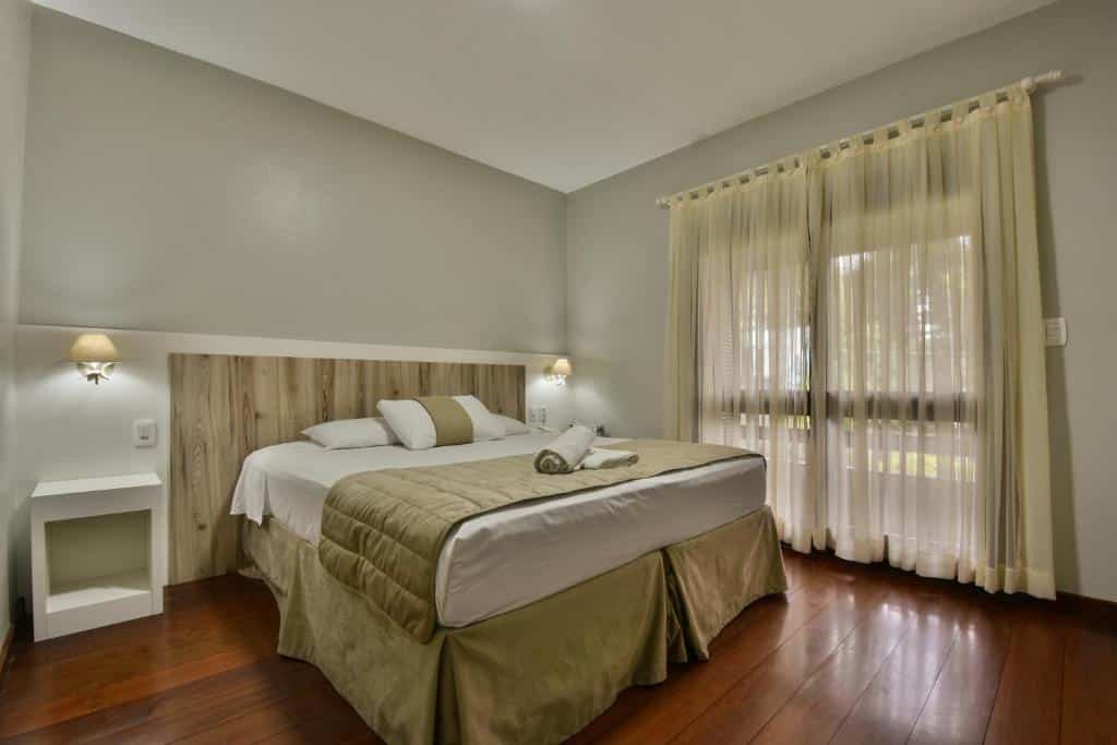Quarto do Hotel Pequeno Bosque. Uma cama de casal do lado esquerdo, com uma cômoda e uma luminária de cada lado. No fundo, a porta da sacada fechada. Foto para ilustrar post sobre hotéis baratos em Gramado.