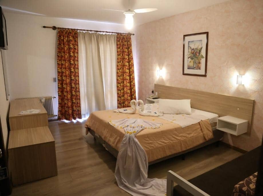 Quarto do Hotel Alpen Hof. Um sofá e uma mesa de casal do lado direito. Do lado esquerdo uma raque e uma televisão. No fundo, cortinas tampando a janela. Foto para ilustrar post sobre hotéis baratos em Gramado.