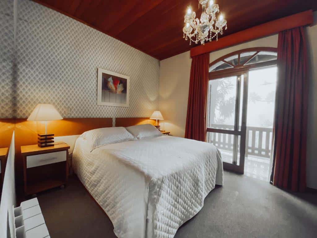 Quarto do Canto Belo Residenziale. Do lado direito uma porta de vidro com a sacada. No lado esquerdo uma cama de casal com uma cômoda e um abajur de cada lado. Foto para ilustrar post sobre hotéis baratos em Gramado.