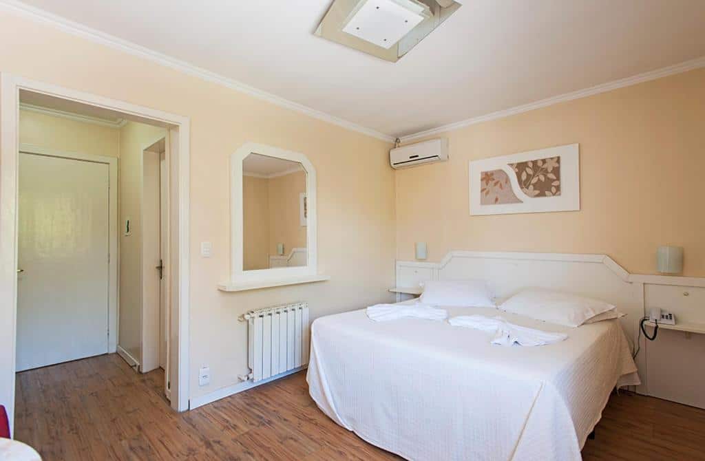 Quarto do Hotel Galo Vermelho. Uma cama de casal do lado direito, com uma prateleira de cada lado, em cima um ar-condicionado. No lado esquerdo, um corredor com a porta do quarto e do banheiro, no meio um espelho. Foto para ilustrar post sobre hotéis baratos em Gramado.