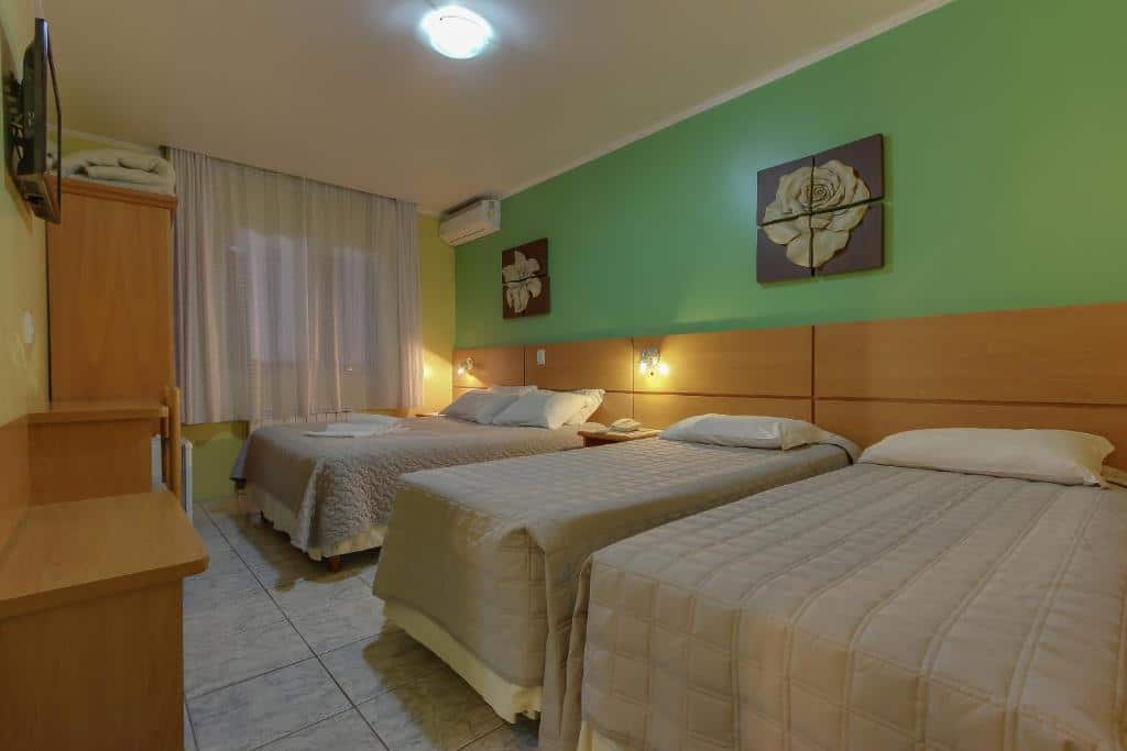 Quarto do Hotel Via Serena. Duas camas de solteiro e uma cama de casal do lado direito, no fundo ar-condicionado. No lado esquerdo, uma mesa de trabalho, um guarda-roupa, frigobar e televisão. Foto para ilustrar post sobre hotéis baratos em Gramado.