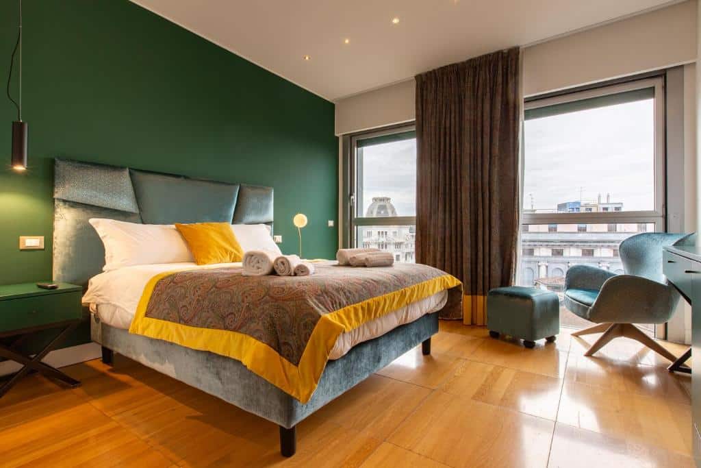 Quarto amplo do Missori Suites com duas janelas grandes com cortinas, uma cama de casal, uma poltrona e um pequeno bufe, há duas mesinhas de cabeceira com luminárias, para representar hotéis baratos em Milão