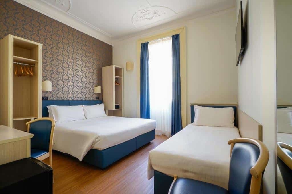 Quarto do Hotel Panizza com uma cama de casal e uma de solteiro, há uma janela com cortinas, uma televisão, chão de madeira, dois pequenos armários de conceito aberto e uma mesa de escritório com uma cadeira