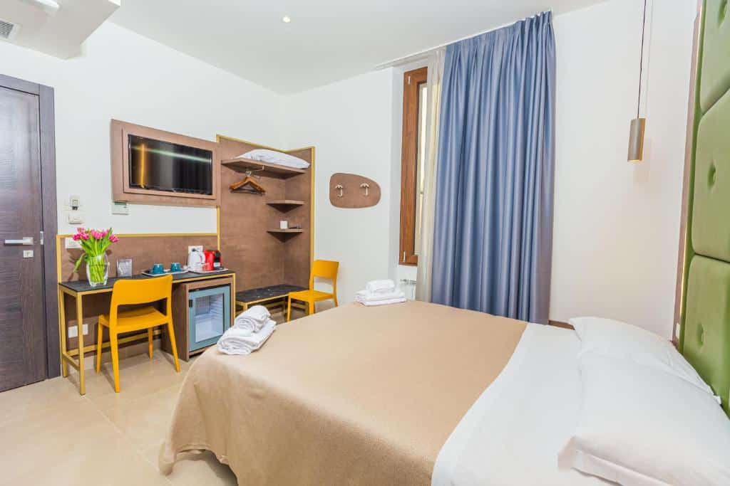 Quarto do Hotel Repubblica com uma janela com cortinas, uma cama de casal, um armário de conceito aberto, uma televisão e uma mesa de escritório com uma cadeira, com um frigobar e alguns itens de cozinha, para representar hotéis baratos em Milão