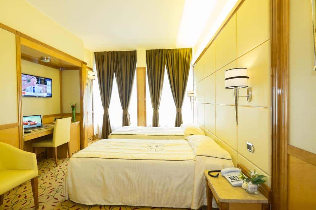 Quarto do Hotel Teco com uma cama de casal e uma de solteiro, além de duas janelas com cortinas, há um pequeno armário, há televisão, mesa de escritório com uma cadeira