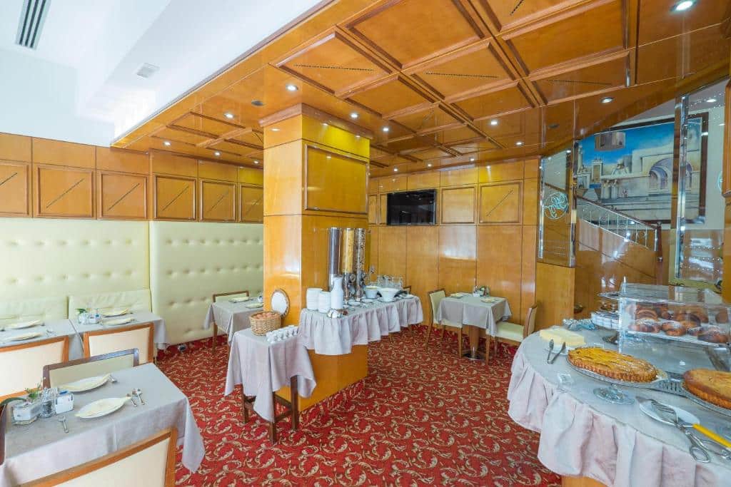 Área de refeições do Hotel Teco com diversas mesas quadradas e cadeiras, carpete vermelho e uma mesa ampla com pães e tortas