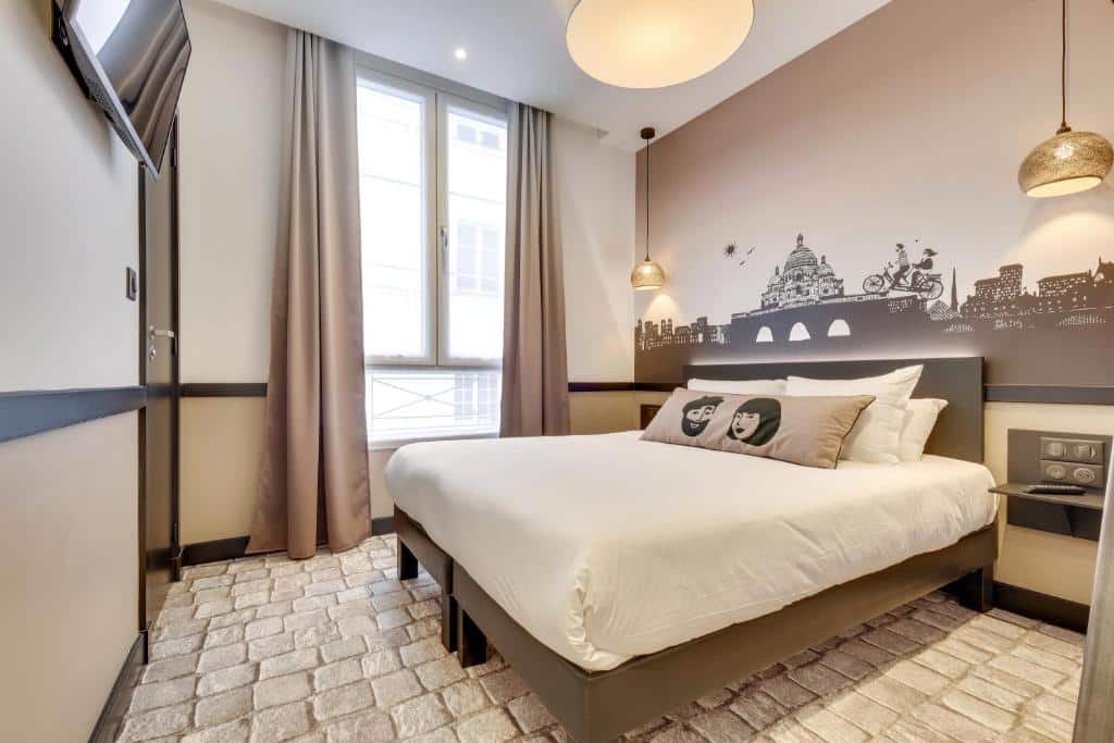 Um pequeno quarto do Hôtel Lucien & Marinette, um dos hotéis baratos em Paris, com uma cama de casal, arte na parede, luzes penduradas, uma janela com cortinas e uma TV na parede.