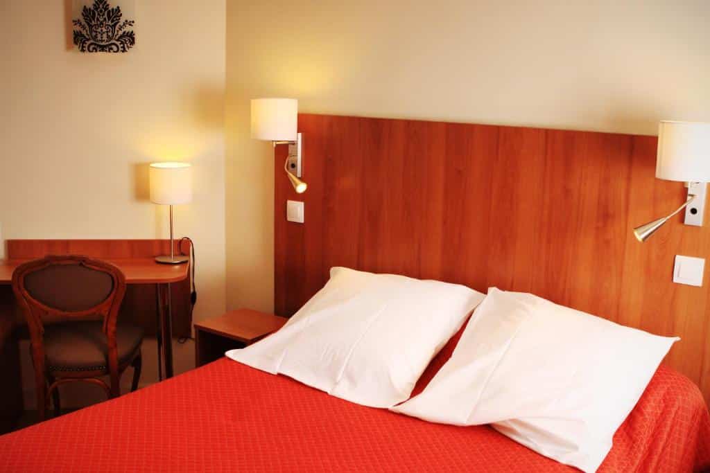 Quarto do Hôtel Marignan com uma cama de casal bem arrumada com travesseiros brancos e uma colcha laranja ao lado de uma escrivaninha de madeira com uma cadeira e uma luminária.
