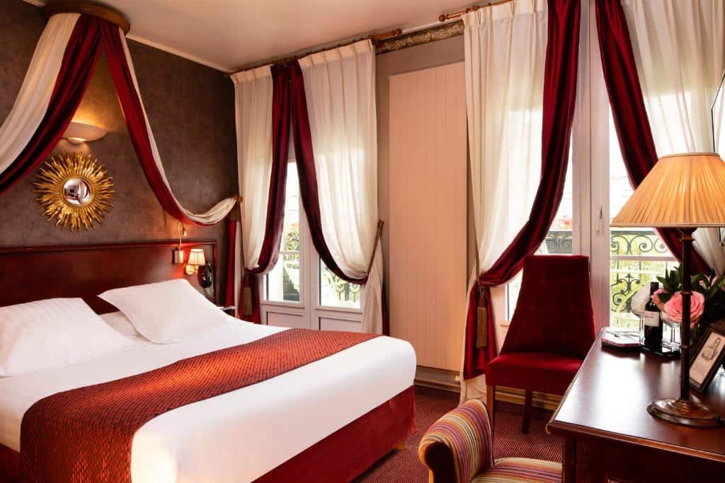 Um quarto do Hotel Britannique, um dos hotéis baratos em Paris, com uma cama de casal, cortinas vermelhas e brancas, uma escrivaninha com uma lâmpada e uma cadeira perto de grandes janelas.