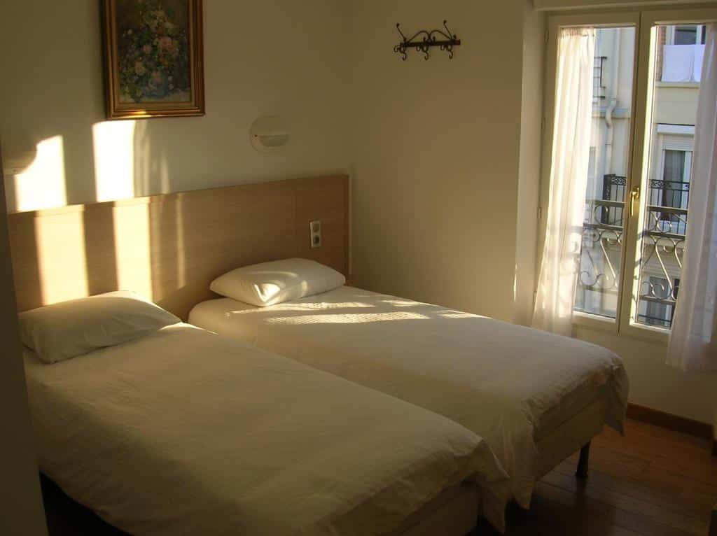 Quarto pequeno do Hotel Darcet com duas camas de solteiro, uma janela com cortinas transparentes, um gancho de parede e uma pintura floral emoldurada.