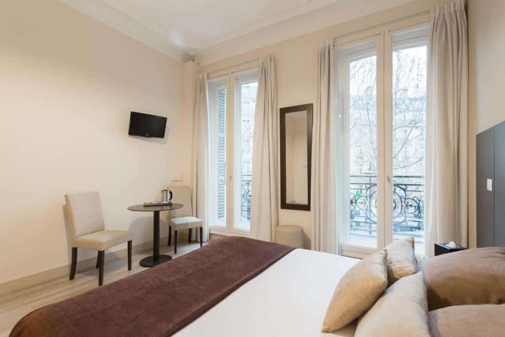 Quarto do Hôtel d’Argenson, um dos hotéis baratos em Paris, com uma cama, duas cadeiras, uma pequena mesa redonda, TV na parede e janelas grandes com cortinas.