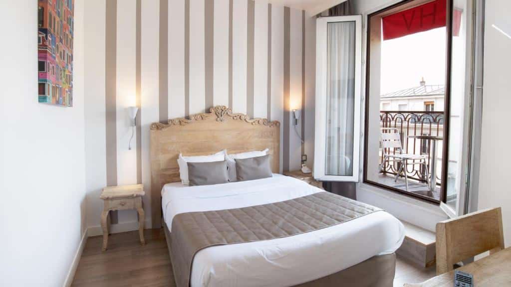Quarto do Hôtel De Venise, um dos hotéis baratos em Paris, com cama de casal, parede com detalhes listrados, mesinhas de cabeceira, janela aberta e varanda com vista para os telhados.