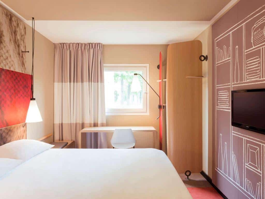 Um quarto do ibis Paris Canal Saint Martin com uma cama, uma escrivaninha e uma cadeira perto de uma janela, uma TV na parede e arte decorativa na parede.