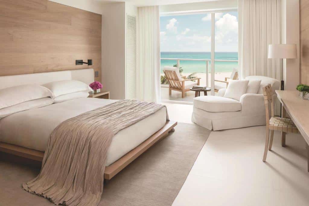 Quarto do The Miami Beach EDITION com cama de casal do lado esquerdo da imagem no centro do quarto do lado esquerdo da cama uma comoda com vaso de flores ao lado em frente a cama uma mesa com cadeira do lado esquerdo da mesa uma poltrona com uma luminária e do lado esquerdo da poltrona uma porta de vidro que dá acesso a varanda com vista para o mar. Representa hotéis boitique em Miami.