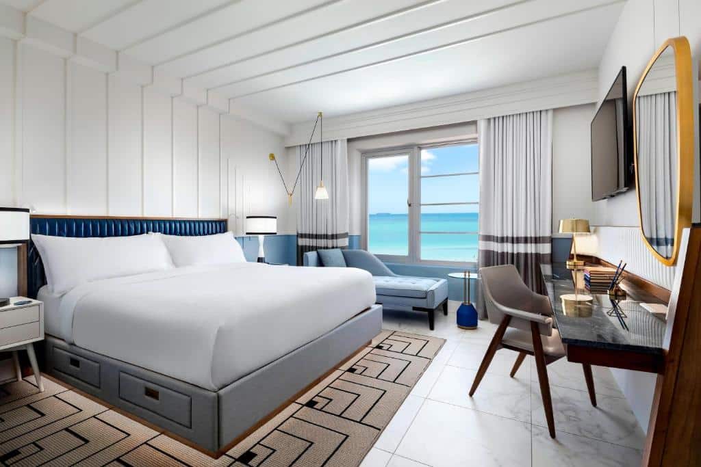 Quarto do Cadillac Hotel & Beach Club, Autograph Collection com cama de casal do lado esquerdo uma cama de casal, em cada lado da cama uma cômoda com luminária em frente a cama uma mesa com cadeira e em cima uma parede com TV presa e do lado esquerdo da cama uma poltrona. Representa hotéis boutique em Miami.