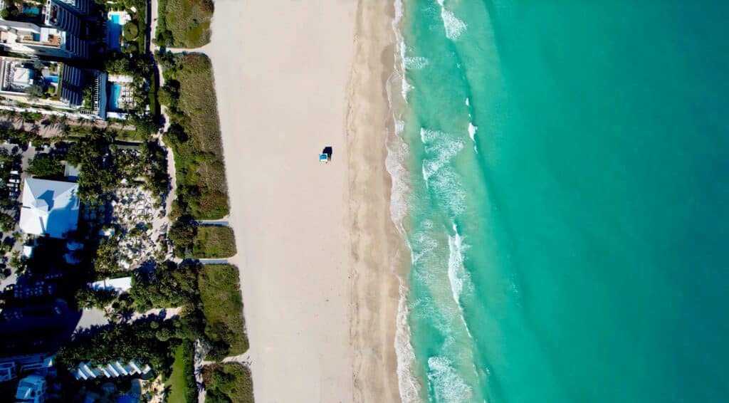 Imagem da praia de Miami, durante o dia com mar do lado direito, faixa de areia no centro e do lado esquerdo casas.