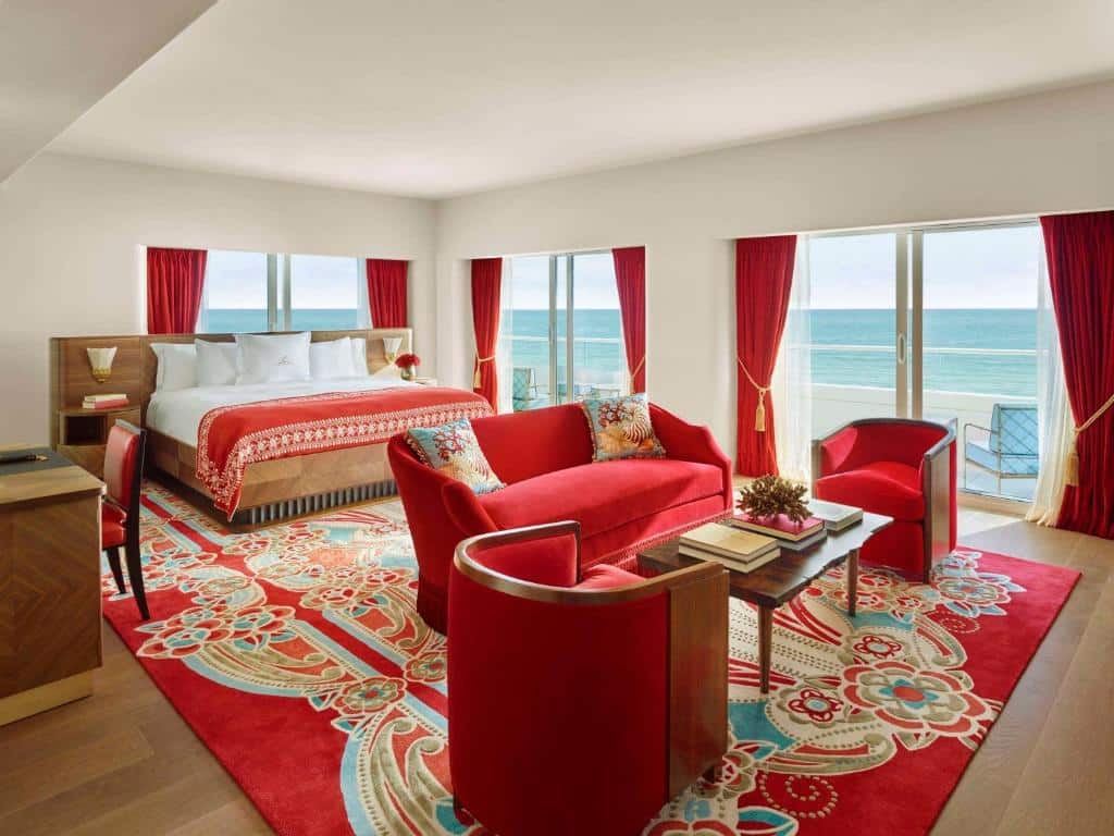 Quarto do Faena Hotel Miami Beach com área de estar a frente com duas poltronas de cada lado no meio uma mesa quadrada e um sofá e ao fundo uma cama de casal e do lado direito portas que dá acesso a praia. Representa hotéis boutique em Miami.