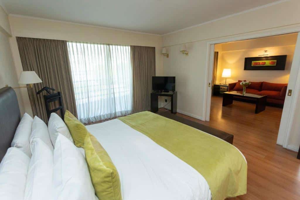 O quarto do Pestana Buenos Aires apresenta uma decoração acolhedora e espaçosa. A imagem mostra uma cama grande e confortável com uma cabeceira estofada em couro escuro. A cama é coberta por lençóis brancos e travesseiros, com uma manta verde e almofadas combinando, acrescentando um toque de cor ao ambiente neutro. À esquerda da cama, há um abajur de pé próximo a uma cadeira preta. As janelas são grandes, com cortinas escuras e uma cortina de tecido translúcido por trás, permitindo a entrada de luz natural enquanto mantém a privacidade. Ao fundo, há uma TV de tela plana posicionada sobre um móvel escuro. Ao lado, uma grande porta de correr se abre para uma sala de estar separada. Este espaço adicional é mobiliado com um sofá vermelho, uma mesa de centro e um abajur, criando um ambiente confortável para relaxar. A decoração é complementada por um quadro de arte na parede, acrescentando um elemento de sofisticação.