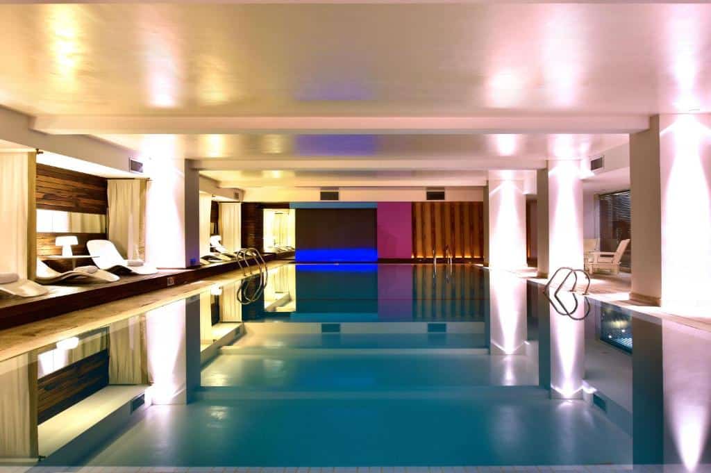 A piscina do hotel Pestana Buenos Aires é uma instalação interna moderna e elegante. O espaço é iluminado por uma luz suave e acolhedora, que reflete na superfície da água cristalina, criando um ambiente tranquilo e relaxante. A piscina é retangular, com bordas bem definidas, e ao lado possui espreguiçadeiras brancas confortáveis, ideais para descansar após um mergulho. As paredes ao redor são decoradas com painéis de madeira e cortinas brancas, que ajudam a criar uma atmosfera intimista. Uma iluminação azul adicional no fundo do espaço adiciona um toque sofisticado e relaxante ao ambiente. As colunas brancas que sustentam o teto são destacadas por uma iluminação indireta, que contribui para o ambiente sereno do local.