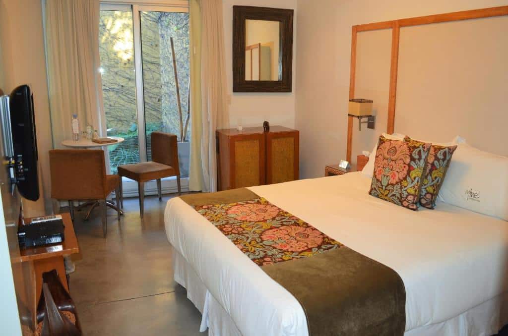 quarto do Mine Hotel com cama de casal e decoração meio vibes com cômoda, tv, espelho de corpo inteiro, mesinha e luminária, além de poltrona e porta de vidro que dá para uma varanda