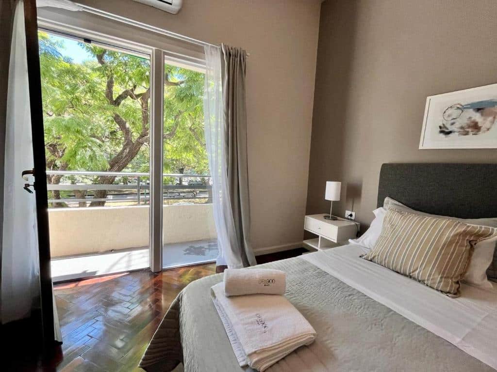 A imagem mostra um quarto de um dos apartamentos do DEL900 em Buenos Aires. O espaço é decorado de forma moderna e aconchegante, com uma paleta de cores neutras que inclui tons de cinza, branco e bege. A cama de casal, central na imagem, possui uma cabeceira estofada em cinza escuro, e está arrumada com lençóis brancos, travesseiros e uma manta bege clara. Sobre a cama, há um conjunto de travesseiros decorativos, incluindo um com listras verticais em bege e branco. Ao lado da cama, uma mesa de cabeceira branca com um design minimalista acomoda uma luminária de cabeceira, proporcionando uma iluminação suave. Há também um ar-condicionado instalado acima da porta de vidro para controle de temperatura. A porta de vidro deslizante dá acesso a uma varanda, permitindo a entrada de luz natural e oferecendo uma vista para uma área arborizada, que confere um ambiente tranquilo e relaxante ao quarto. As cortinas brancas translúcidas, ao lado da porta, adicionam um toque leve e elegante ao espaço.