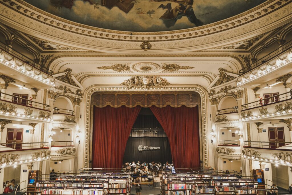 A imagem mostra o interior da Livraria El Ateneo, localizada em Buenos Aires, Argentina. O local originalmente era um teatro, e mantém a grandiosidade e a elegância arquitetônica de sua época. O teto é adornado com um afresco detalhado, e as varandas laterais possuem ornamentos dourados e iluminados que destacam a decoração clássica.
No centro da imagem, há um grande palco com uma cortina vermelha imponente, que remete aos tempos em que ali eram realizadas apresentações teatrais. A área onde antes ficava a plateia agora está repleta de estantes e livros, com várias pessoas explorando o espaço. A atmosfera combina o esplendor histórico do teatro com a tranquilidade e o encanto de uma grande livraria.