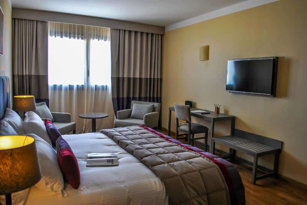 quarto do Hotel Grand Brizo Buenos Aires com cama de casal, tv e mesa com cadeira de escritório em frente, além de um apoio para mala, do outro lado há mais uma mesinha com duas poltronas, janela grande e cortinas
