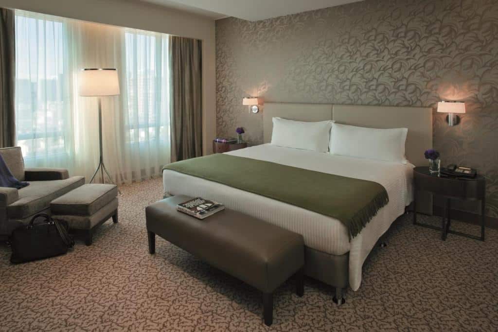 O quarto do Alvear Art Hotel é decorado em tons neutros, com uma cama king-size central, coberta por uma colcha branca e um cobertor verde. Ao lado da cama, há uma poltrona confortável com um apoio para os pés e uma luminária de chão ao lado, proporcionando um espaço de leitura. Na frente da cama, há uma banqueta estofada e mesas de cabeceira com abajures modernos. As paredes são decoradas com um padrão discreto de folhas, e uma grande janela com cortinas permite a entrada de luz natural, criando um ambiente acolhedor e sofisticado.