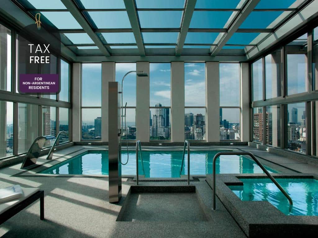 A imagem retrata a piscina coberta do Alvear Art Hotel, um dos hotéis com piscina em Buenos Aires localizada em um andar alto com janelas amplas que proporcionam uma vista panorâmica impressionante da cidade de Buenos Aires. A área da piscina é sofisticada, com um design moderno e elegante. As paredes de vidro permitem uma abundante entrada de luz natural e oferecem uma vista deslumbrante dos arranha-céus e da paisagem urbana ao redor. A piscina tem bordas de granito cinza, combinando com o piso e as estruturas metálicas que sustentam o teto de vidro. Há um espaço de banheira de hidromassagem ao lado da piscina, também revestido de granito, que complementa a área de relaxamento. As espreguiçadeiras são colocadas próximas às janelas, oferecendo aos hóspedes um lugar confortável para descansar enquanto desfrutam da vista.