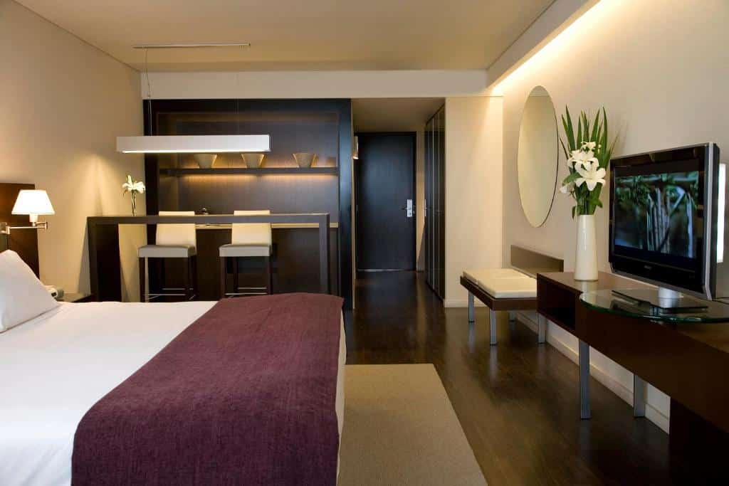 quarto do Hotel Madero Buenos Aires com cama de casal, mesa e cadeira em frente, além de uma tv e, ao lado, uma mesa comprida e duas cadeiras altas com luminária acima