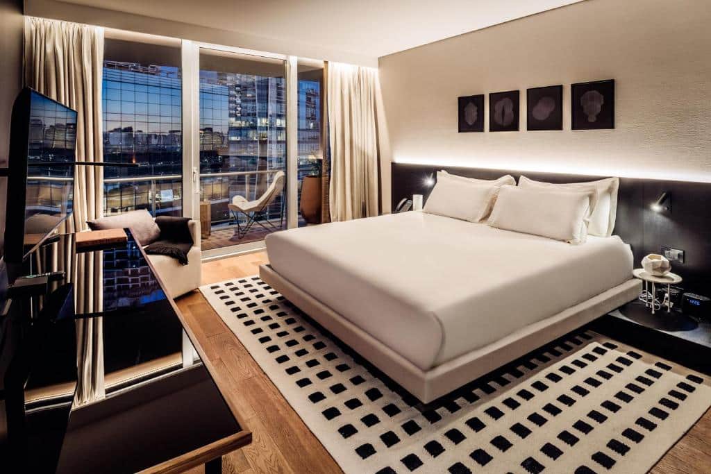 quarto amplo e com decoração moderna do SLS Buenos Aires Puerto Madero com cama de casal grande, mesinha e luminária de ambos os lados, tv e escrivaninha em frente, com poltrona e janela grande com varanda em que há duas outras cadeiras e uma mesinha com vista, o chão está decorado com um padrão quadriculado em branco com quadradinhos pretos, há quadros na parede
