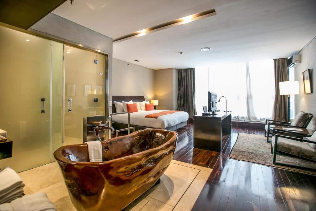 A imagem mostra um quarto de hotel do Mio Buenos Aires, que é elegante e moderno. O quarto possui uma cama grande com cabeceira estofada em tons de cinza. No centro do quarto, destaca-se uma banheira autônoma em acabamento de madeira polida, dando um ar sofisticado e natural ao ambiente. A banheira é acompanhada de uma torneira moderna e toalhas dispostas ao lado. A parede de vidro fosco ao fundo separa o banheiro, que contém uma área de chuveiro visível. O piso de madeira escura combina com as cortinas pesadas que emolduram as janelas, permitindo a entrada de luz natural que ilumina todo o espaço. A frente da cama, há uma televisão sobre uma mesa preta, e ao lado, duas poltronas confortáveis com uma mesa de centro sobre um tapete.
