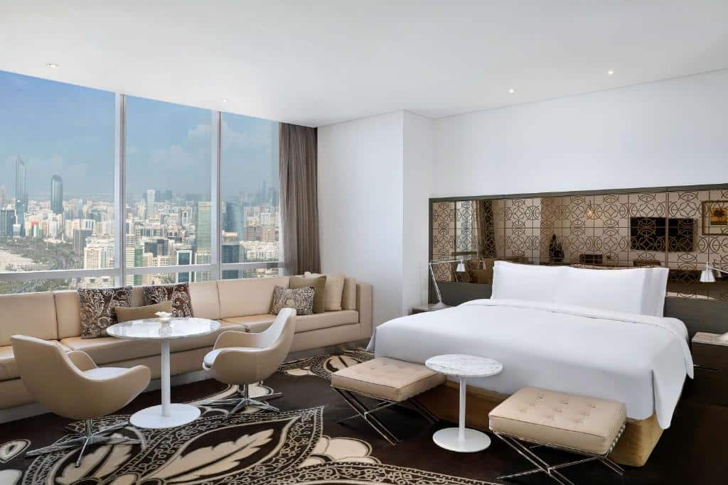 Quarto do Conrad Abu Dhabi Etihad Towers, no lado direito uma cama de casal, com um abajur de cada lado, na frente dois bancos estufados e uma mesinha. No lado esquerdo um sofá, uma mesa com duas cadeiras, e uma parede de vidro com vista para a cidade. 
