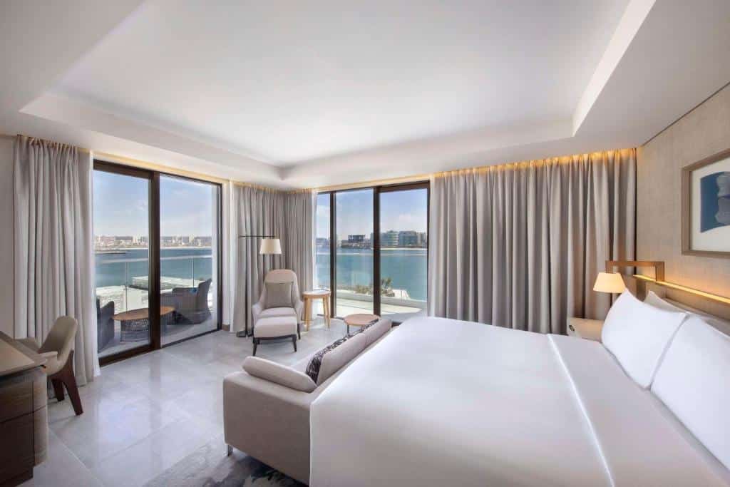 Quarto do Hilton Abu Dhabi Yas Island. Uma cama de casal no lado direito com um abajur atrás. Na frente da cama um sofá, do lado esquerdo uma mesa de trabalho e no lado direito uma poltrona com um abajur. Nas paredes do quarto de vidro, cortinas e uma sacada com vista para o mar. 