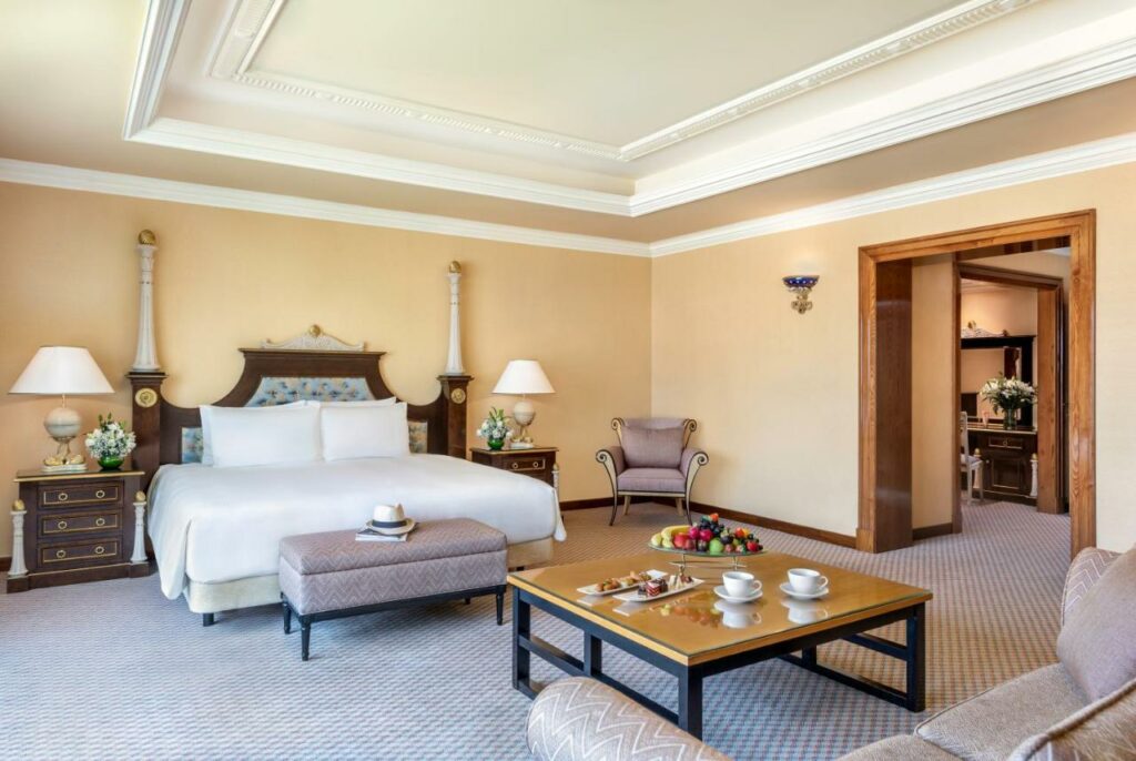 Quarto do Al Raha Beach Hotel. Um sofá no canto direito com uma mesa quadrada no centro. De frente, um sofá com uma cama atrás de casal. De cada lado da cama uma cômoda com abajur. Do lado direito da cama, uma poltrona. No lado direito do quarto o corredor da entrada.