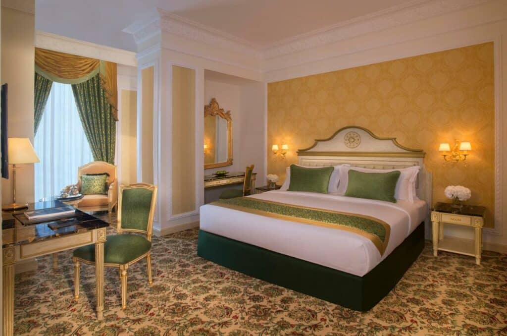 Quarto do Royal Rose Hotel. Uma cama de casal no lado direito com um criado mudo de cada lado e um abajur. Uma penteadeira com um espelho do lado esquerdo da cama. De frente para a cama uma mesa de trabalho e no fundo uma poltrona com cortinas na janela.
