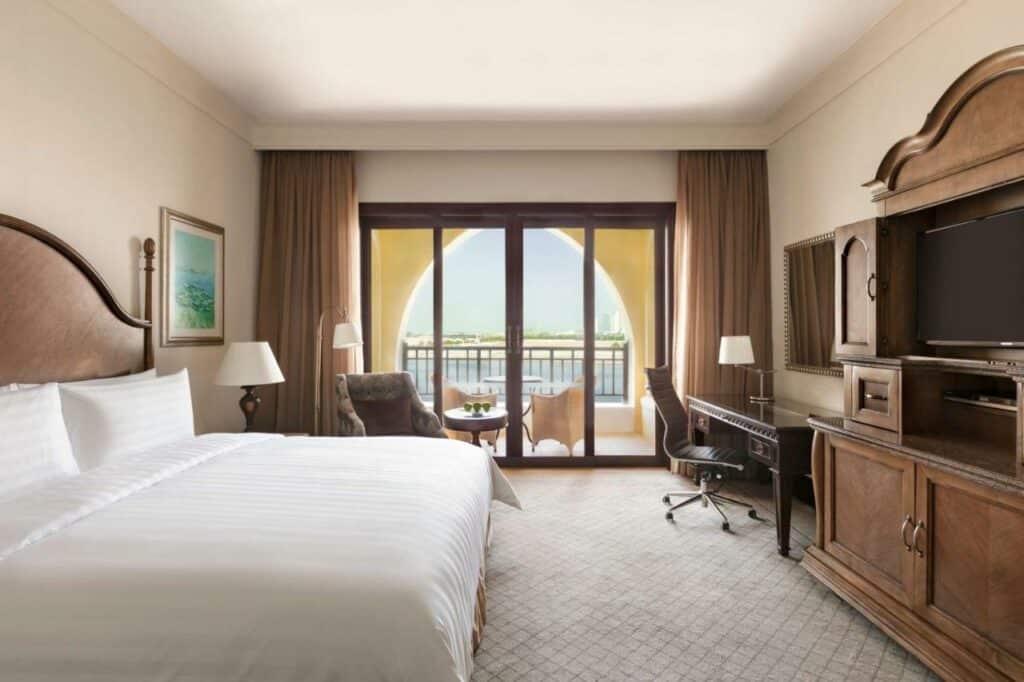 Quarto do Shangri-La Qaryat Al Beri, Abu Dhabi. Uma cama de casal no lado esquerdo, atrás dois abajures e uma poltrona comuma mesinha redonda. De frente para a cama, uma cômoda com uma televisão, e do lado uma mesa de trabalho com um espelho. No fundo, a varanda do quarto com duas cadeiras e uma mesa e a vista para o mar.