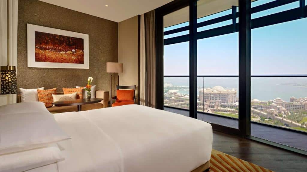 Quarto do Grand Hyatt Abu Dhabi Hotel & Residences Emirates Pearl. Uma cama de casal no lado esquerdo, atrás um sofá, com dois abajures, uma mesa redonda, uma poltrona e um quadro. De frente para a cama, uma parede de vidro com uma sacada e vista para a cidade e o mar. Foto para ilustrar post sobre Hotéis em Abu Dhabi.