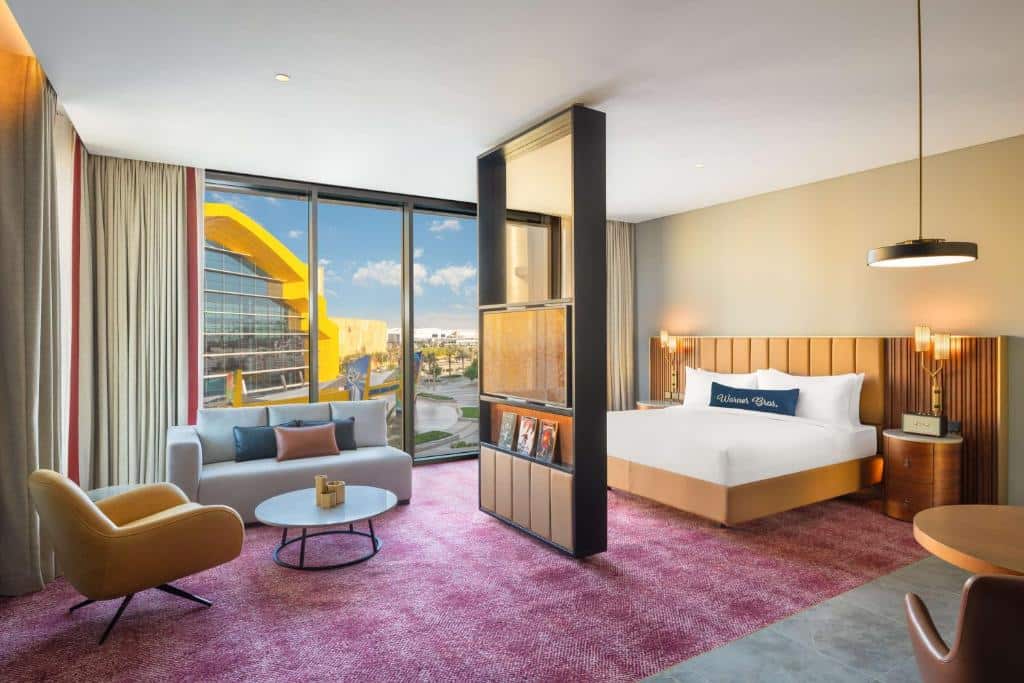 Quarto do The WB Abu Dhabi, Curio Collection By Hilton, com uma poltrona no lado esquerdo com uma mesa de centro e um sofá. No lado direito uma mesinha com cadeira, uma cama de casal com uma cômoda e um abajur de cada lado. No meio do quarto, dividindo, um painel de madeira com uma televisão. No fundo uma parede de vidro com vista para a entrada do hotel. 