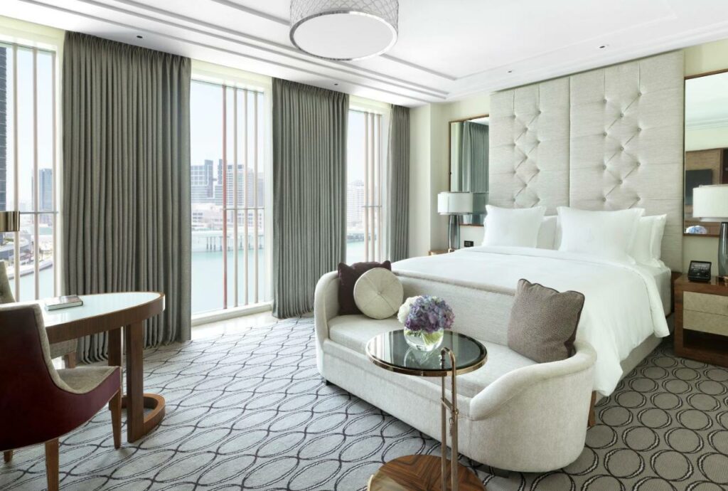 Quarto do Four Seasons Hotel Abu Dhabi at Al Maryah Island, com uma cama de casal no lado direito com um abajur de cada lado e na frente um sofá com uma mesinha de vidro. No lado esquerdo uma mesa com duas cadeiras e atrás uma parede de vidro com cortinas e vista para a cidade. 